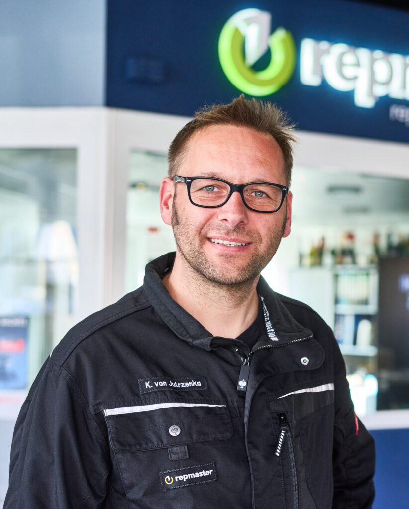 Ein Mitarbeiter steht im Eingangsbereich eines repmaster‑Autohauses und trägt eine schwarze Arbeitsjacke mit Namensschild und Firmenlogo. Im Hintergrund sind Glaswände, Regale und das beleuchtete repmaster‑Logo zu sehen.
