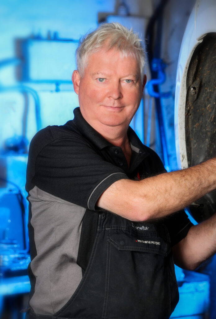 Porträt einer Person in einer Autowerkstatt mit hellem, kurzem Haar und dunkler Arbeitskleidung. Der Mann trägt eine schwarze Latzhose über einem schwarzen Polo mit grauen Einsätzen. Die Person arbeitet am Radkasten eines angehobenen Fahrzeugs; im Hintergrund leuchten blaue Werkstattelemente.