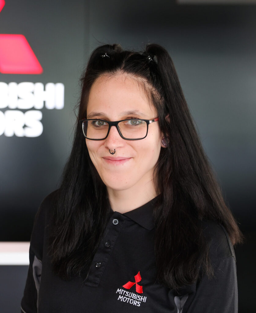 Porträt einer lächelnden Person mit schwarzer Brille, Septum-Piercing und halb hochgebundenem dunklem Haar. Sie trägt ein schwarzes Polo mit rotem Mitsubishi‑Motors‑Logo. Im unscharfen Hintergrund ist ein Autohaus‑/Servicebereich mit Teile des Markenzeichens zu sehen.