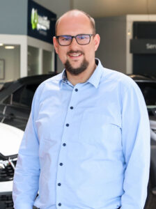 Porträt einer Person in einem Autohaus‑Showroom. Der Mann trägt ein hellblaues Hemd mit dunklen Knöpfen und steht zwischen Fahrzeugen. Im Hintergrund sind ein „Service“-Schild und ein repmaster‑Logo zu erkennen.