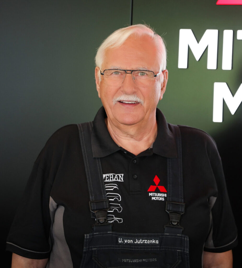 Porträt eines lächelnden älteren Werkstattmitarbeiters mit weißem Haar, Brille und Schnurrbart. Er trägt eine dunkle Latzhose über einem schwarzen Polo mit rotem Mitsubishi‑Motors‑Logo. Im unscharfen Hintergrund ist ein Autohaus‑/Servicebereich mit Teilen des Markenzeichens zu sehen.