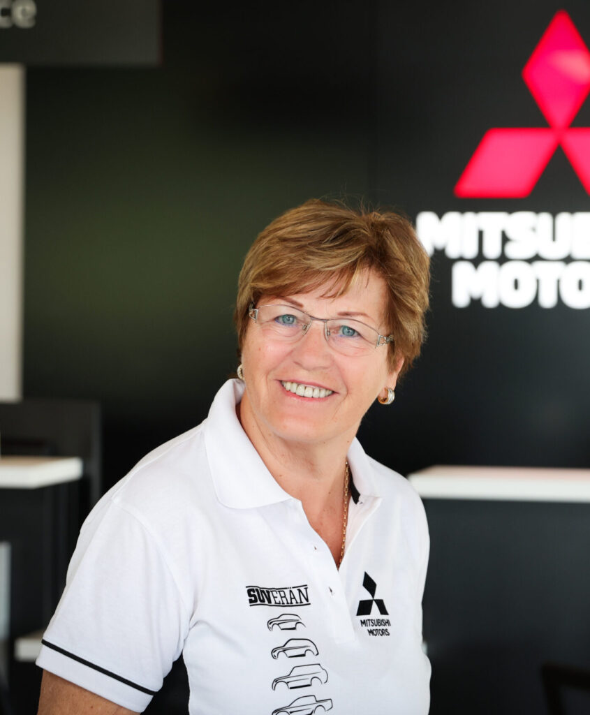 Porträt einer lächelnden Frau in einem Autohaus‑Servicebereich. Die Person trägt ein weißes Poloshirt mit Mitsubishi Motors‑Logo und grafischen Autokonturen. Im Hintergrund sind unscharf das rote Markenlogo und Teile der Einrichtung zu sehen.