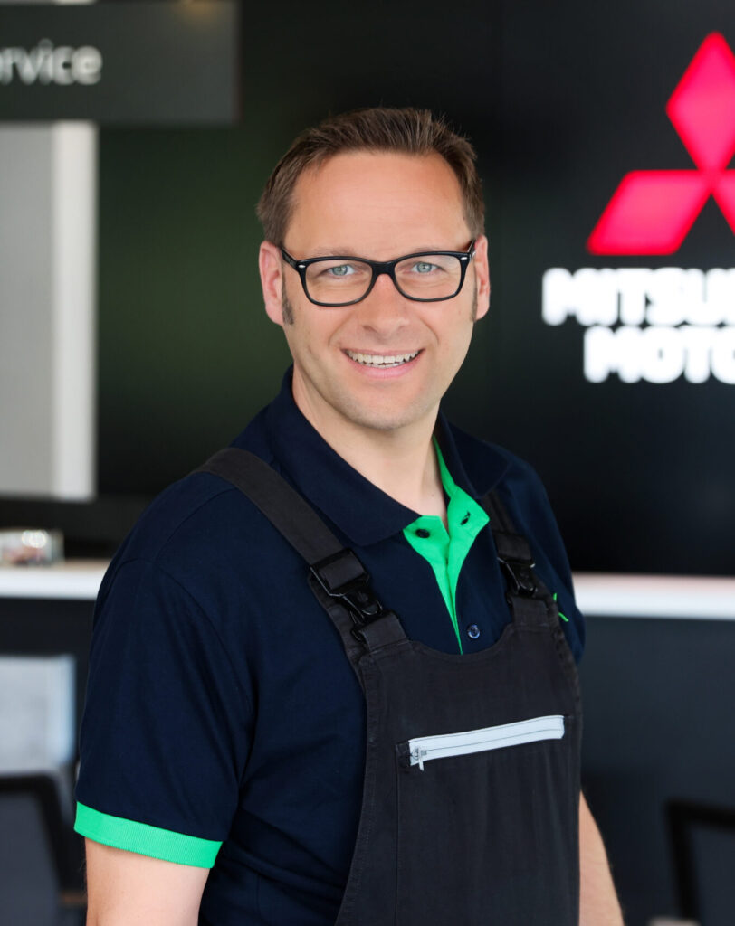 Porträt eines lächelnden Werkstattmitarbeiters mit schwarzer Brille in dunkler Latzhose und navyfarbenem Polo mit grünem Kragen. Er steht in einem Autohaus‑ bzw. Servicebereich und blickt in die Kamera. Im Hintergrund sind unscharf ein rotes Markenlogo und ein Schild mit der Aufschrift „Service“ zu sehen.