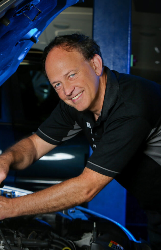 Eine Person arbeitet in einer Autowerkstatt am geöffneten Motorraum. Er trägt ein dunkles Poloshirt und beugt sich unter die hochgeklappte Motorhaube. Im Hintergrund ist die Werkstattausstattung zu erkennen.