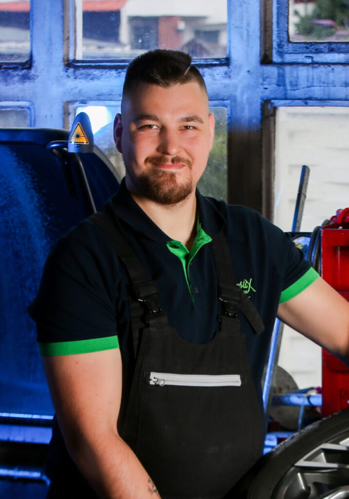 Porträt eines lächelnden Werkstattmitarbeiters in schwarzer Latzhose und dunkelblauem Polo mit grünem Kragen. Er steht in einer Kfz‑Werkstatt neben einem Rad/Reifen, im Hintergrund sind Werkzeuge und eine rote Maschine zu sehen. Blaues Licht fällt durch die Fenster, ein gelbes Warnsymbol ist an einem Gerät erkennbar.