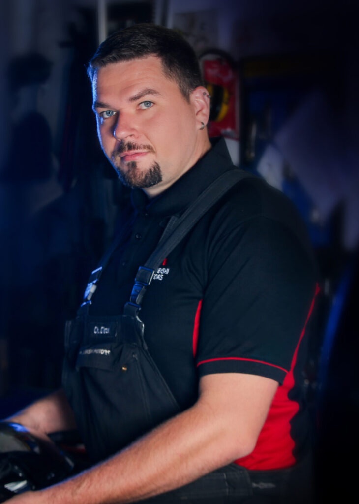 Porträt einer Person in einer Werkstatt. Er trägt eine schwarze Latzhose und ein schwarzes Polohemd mit roten Einsätzen und kleinem Logo. Die Person hält ein Teil in den Händen, der Hintergrund zeigt unscharfe Werkstattausrüstung.